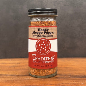 A glass jar of Tradition Spice Company Honey Aleppo Pepper Dry Rub Seasoning, featuring a black metal lid and a red and white label, set on a wooden surface against a dark gray background.