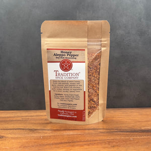 A stand-up pouch of Tradition Spice Company Honey Aleppo Pepper dry rub seasoning, displayed on a wooden surface against a dark gray background.