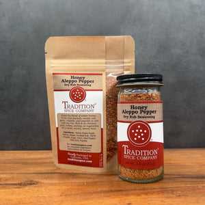 A kraft paper pouch and a clear glass jar of Tradition Spice Company Honey Aleppo Pepper Dry Rub Seasoning, displayed on a wooden surface against a dark gray background.