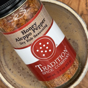 A close-up of a spice jar labeled 'Honey Aleppo Pepper Dry Rub Seasoning' by Tradition Spice Company, resting inside a ceramic bowl.