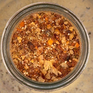 A top-down view of a clear glass jar filled with a textured, granular brown and orange mixture resembling bark, wood chips, or a natural spice blend, set against a speckled beige countertop.