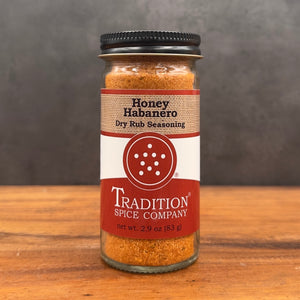 A glass jar of Tradition Spice Company Honey Habanero Dry Rub Seasoning sits on a wooden surface against a dark gray background.
