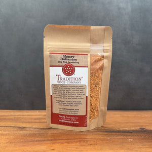 A stand-up kraft paper pouch of Tradition Spice Company Honey Habanero Dry Rub Seasoning, standing on a wooden surface against a dark gray background.