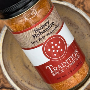 A close-up of a spice rub seasoning bottle labeled 'Honey Habanero Dry Rub Seasoning' by Tradition Spice Company, resting inside a rustic ceramic bowl.