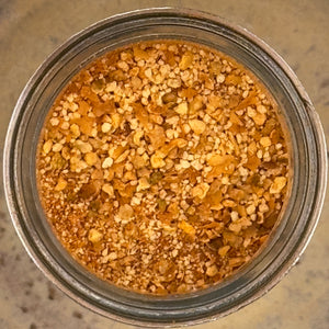Top-down view of a clear glass jar filled with a granola-like mixture of oats, nuts, seeds, and dried herbs on a speckled countertop.