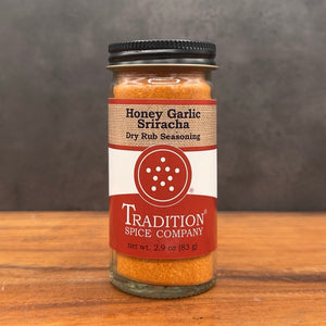 A glass jar of Tradition Spice Company Honey Garlic Sriracha Dry Rub Seasoning, filled with brown seasoning and topped with a black lid, set on a wooden surface against a dark gray background.
