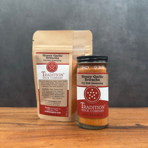 A stand-up pouch and a glass jar of Tradition Spice Company Honey Garlic Sriracha dry rub seasoning, displayed on a wooden surface against a dark gray background.