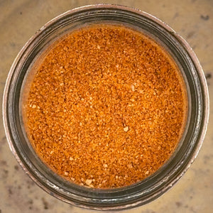A top-down view of a clear glass jar filled with a fine brown-orange spice or seasoning blend, set on a speckled beige countertop.
