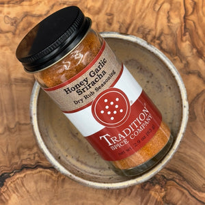 A bottle of Tradition Spice Company Honey Garlic Sfrachta dry rub seasoning, placed diagonally inside a rustic ceramic bowl on a textured wood background.