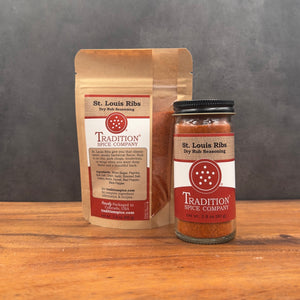 A glass jar and a stand-up pouch of Tradition Spice Company St. Louis Ribs Dry Rub Seasoning, displayed side-by-side on a wooden surface against a dark gray background.