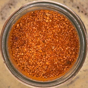 Top-down view of a clear glass jar filled with a coarse brown spice or seasoning blend on a light speckled countertop.