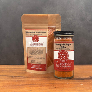 A stand-up pouch and a glass jar of Tradition Spice Company Memphis Style Ribs Dry Rub Seasoning displayed on a wooden surface against a dark gray background.