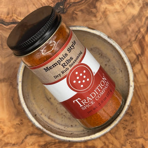 A glass jar of Tradition Spice Company Memphis Style Dry Rub Seasoning with a black lid, resting on a rustic ceramic dish over a wood grain surface.