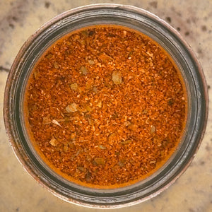 Top-down view of a spice blend inside a clear glass jar, showing a textured mix of orange, brown, and reddish seasoning on a speckled beige countertop.