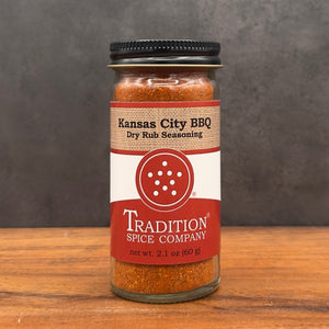 A glass jar of Tradition Spice Company Kansas City BBQ Dry Rub Seasoning sits on a wooden surface against a dark gray textured background.