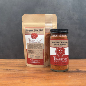 A jar and a stand-up pouch of Tradition Spice Company Kansas City BBQ Dry Rub Seasoning displayed on a wooden surface against a dark gray background.