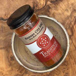 A bottle of Tradition Spice Company Kansas City BBQ Dry Rub Seasoning resting inside a small rustic ceramic dish on a wood grain surface.