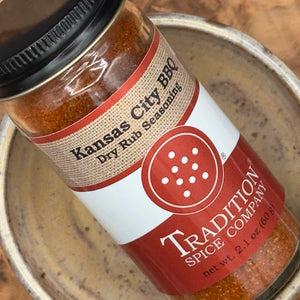 A close-up of a glass jar of Kansas City BBQ Dry Rub Seasoning by Tradition Spice Company, showing the spice rub inside and the label details, resting in a metal bowl.