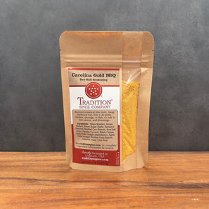 A stand-up pouch of Tradition Spice Company Carolina Gold BBQ dry rub seasoning, displayed on a wooden surface against a dark gray background.
