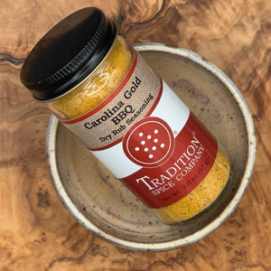 A bottle of Tradition Spice Company Carolina Gold BBQ Dry Rub Seasoning resting inside a small rustic ceramic bowl on a textured wood surface.