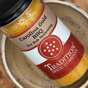A close-up of a spice jar labeled Carolina Gold BBQ Dry Rub Seasoning by Tradition Spice Company, resting inside a metal bowl.