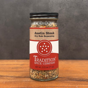A glass jar of Austin Steak Dry Rub Seasoning by Tradition Spice Company, featuring a black lid and a label with red, white, and tan sections, set on a wooden surface against a dark gray background.