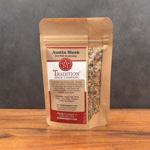 A stand-up pouch of Tradition Spice Company Austin Steak Dry Rub Seasoning, featuring a kraft paper design with a clear window showing the seasoning blend inside, set on a wooden surface against a dark gray background.