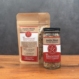 A kraft paper pouch and a clear glass jar of Tradition Spice Company Austin Steak Dry Rub Seasoning, displayed on a wooden surface against a dark gray background.