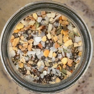 A top-down view of a clear glass jar filled with a multicolored salt mixture containing various crystals, flakes, and small pieces of dried herbs or spices.