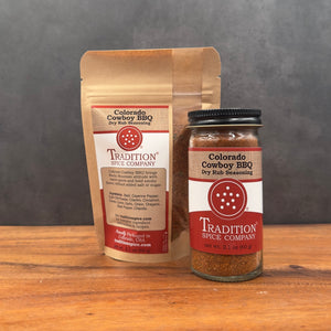 A glass jar and a stand-up pouch of Tradition Cowboy BBQ Dry Rub Seasoning from Tradition Spice Company, displayed on a wooden surface against a dark gray background.