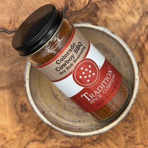 A glass jar of Tradition Spice Company Colorado Cowboy BBQ Dry Rub Seasoning, featuring a black lid and a label with red, white, and burlap textures, resting on a rustic ceramic plate over a wood grain surface.