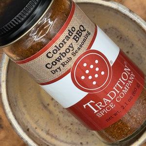 A close-up of a glass jar of Tradition Spice Company Colorado Cowboy BBQ Dry Rub Seasoning resting inside a rustic metal bowl.