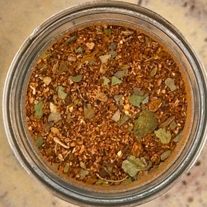 A top-down view of a spice blend inside a clear glass jar, showing a mixture of brown ground spices with dried green and beige herbs.