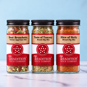 Three glass jars of Tradition Spice Company bread dips lined up on a white marble surface against a light blue background. The jars contain Italian appetizer mix, bread dip mix, and sliced of icily bread dip mix, with black lids and red and white labels.