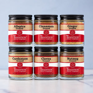 Six glass jars of Tradition Spice Company ground spices arranged in two rows of three on a light marble surface against a pale blue-gray background. The jars include Allspice, Cinnamon, Ginger, Cardamom, Cloves, and Nutmeg, each with a black lid and a red-and-beige label.