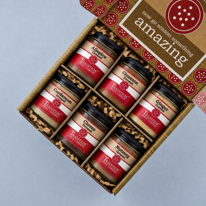 Six glass jars of Tradition Spice Company ground spices arranged in two rows of three in a Tradition gift box. The jars include Allspice, Cinnamon, Ginger, Cardamom, Cloves, and Nutmeg, each with a black lid and a red-and-beige label.