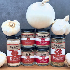 Six spice jars from the Garlic & Onion Cookhouse Kit are arranged in two rows amid garlic bulbs and a large white onion, set against a gray background.