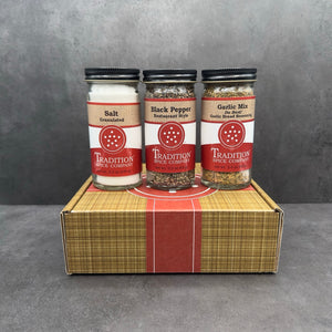 The Welcome Home Gift Box, featuring Salt, Black Pepper, and Garlic Mix jars with the Tradition Spice Company’s red logo, sits on a brown striped box. The gray background adds elegance—an ideal housewarming gift.