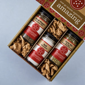 A gift box of four spice blends from Tradition Spice Company, including Black Pepper, Salt, and Garlic Mix, arranged in a cardboard tray with brown paper shred on a light gray background.