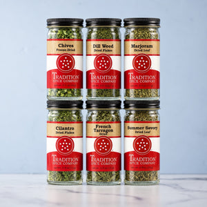 Six glass spice jars from Tradition Spice Company arranged in two rows of three on a light marble surface against a pale blue background. The jars contain various dried herbs and spices including Chives, Dill Weed, Marjoram, Cilantro, French Tarragon, and Summer Savory, each with a black lid and a red, white, and beige label.