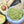 A gray marble bowl filled with guacamole made using the Guac Me Up! Guacamole Maker’s Kit sits on a wooden surface, with a tortilla chip dipped in it. Halved avocados and more chips are visible in the background.