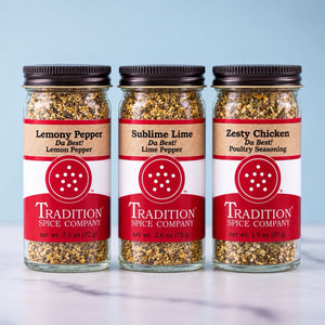 Three glass jars of Tradition Spice Company seasoning blends lined up on a light marble surface against a pale blue background. The jars are labeled Lemony Pepper, Sublime Lime, and Zesty Chicken.