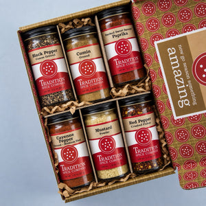 A gift box of six spice jars from Tradition Spice Company, arranged in a brown kraft box with shredded paper filler. The open lid on the right shows a patterned interior and a label that reads 'amazing' with 'now 90 season spices.
