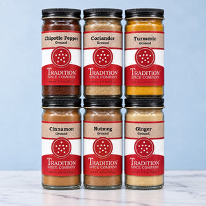 Six glass jars of ground spices from Tradition Spice Company are arranged in a 2x3 grid. The spices include Chipotle Pepper, Coriander, Turmeric, Cinnamon, Nutmeg, and Ginger.