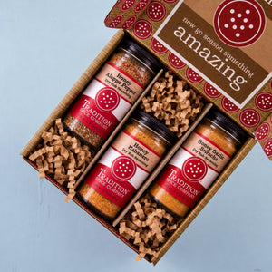 A gift box of three spice jars from Tradition Spice Company, displayed diagonally on a light blue background. The open kraft box contains three clear jars of honey-infused spices (Masa Pepper, Habanero, Garlic Rub) nestled in brown crinkle paper, with the lid showing a 'now go season something amazing' label.