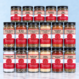 A 12-pack assortment of glass spice jars from Tradition Spice Company, arranged in a grid on a light gray background. The jars contain various ground and dried spices such as basil, parsley, thyme, rosemary, sage, zesty chicken, mi taco, italian, steak & burger, seafood, chili & stew, kosher salt, black pepper, garlic salt, onion, paprika, and oregano.