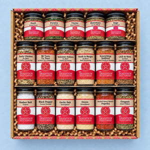 A gift box of 16 jars of Tradition Spice Company whole spices, arranged in four rows of four on a light blue background.