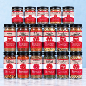 A 16-pack assortment of Tradition Spice Company whole spice blends in clear glass jars with black lids and red-and-white labels, arranged in a grid on a light marble surface against a pale blue background.