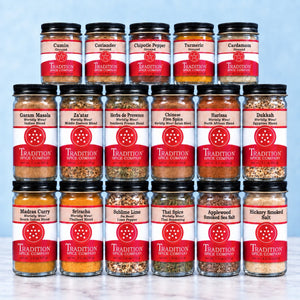 A 16-pack assortment of Tradition Spice Company gourmet spice jars arranged in a 4x4 grid. The clear glass jars reveal colorful ground spices inside, each topped with a black lid and a red and white label showing the brand name and variety such as Cumin, Coriander, Chipotle Pepper, Turmeric, Cardamom, Garum Masala, Za'atar, Herb de Provenches, Chinese Fire Spice, Harlessa, Dukhla, Madras Curry, Srinivas, Sublime Lime, Thai Spice, Applewood Sanded Sea Salt, and Hickory Smoked Salt.