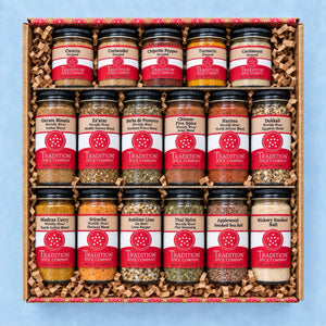 A gift box of 20 jars of Tradition Spice Company hot and spicy blends, arranged in a wooden tray with crinkle paper packing material, set against a light blue background.